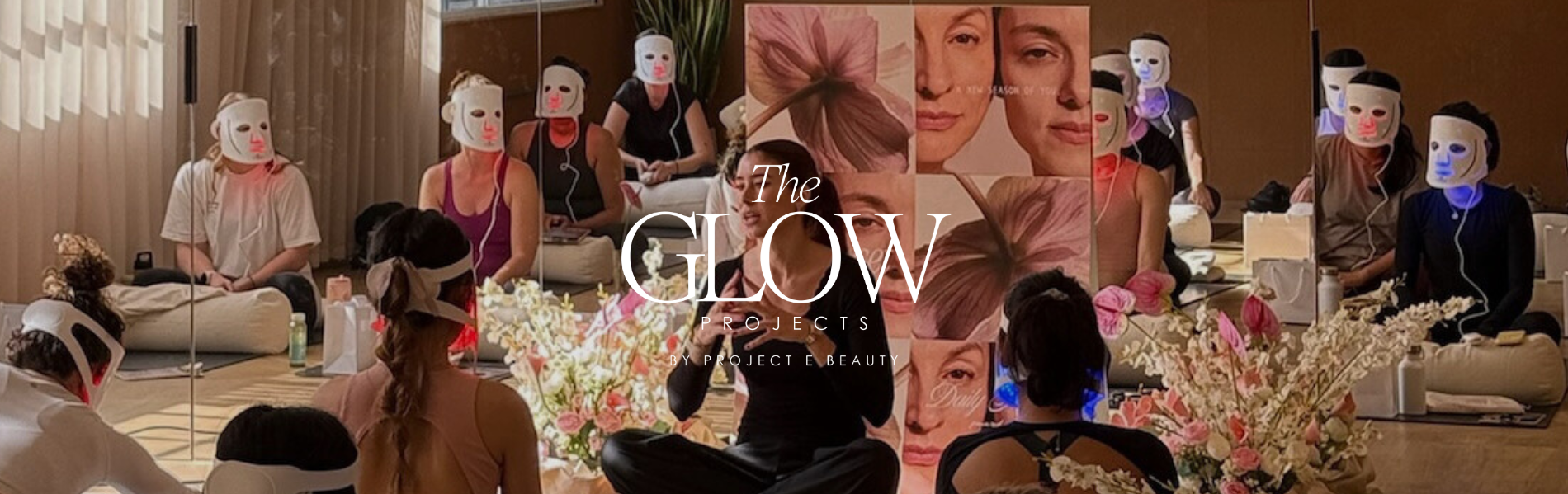 Group wellness session with people wearing glowing Project E Beauty LumaLux Face LED Red light therapy masks, sitting on yoga mats in a studio decorated with flowers and skincare posters.